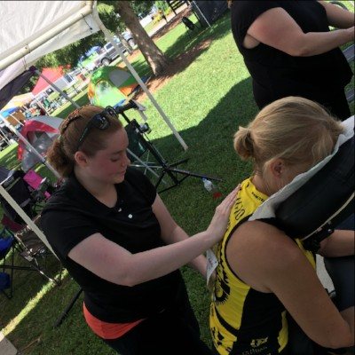 Chair massage under a tent at an outdoor event, Emma working on a seated client's shoulders