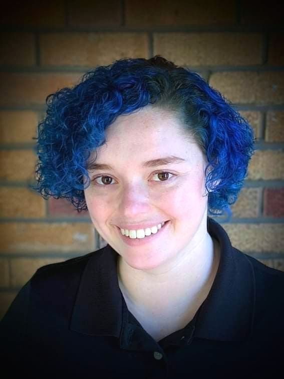 Emma Bufalini, founder of KinesioCal, a massage therapist with short curly blue hair, smiling in a black polo in front of a brick wall